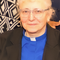 Portrait of an older woman in a blue clerical shirt and black jacket.