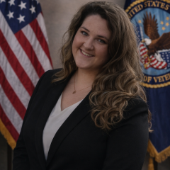 A woman in a black blazer and white shirt smiles, standing in front of American and Veterans Affairs flags.