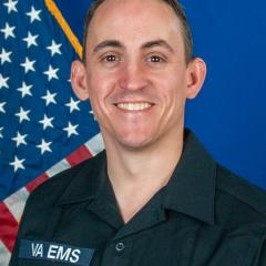 Man in black VA EMS uniform smiles in front of American flag.