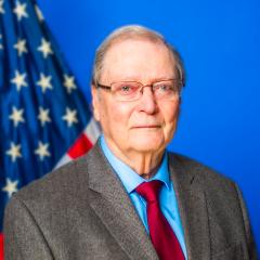 Older man in suit and red tie stands in front of American flag.