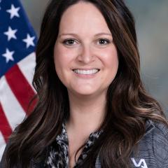 Woman with long brown hair smiling in front of American flag.