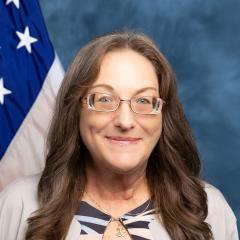 Woman with glasses and a gray jacket smiling in front of an American flag.