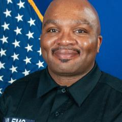 Man in black uniform with "VA EMS" patch smiling in front of a US flag.