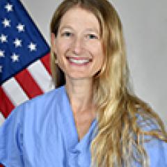 Smiling blonde woman in blue scrubs in front of an American flag.
