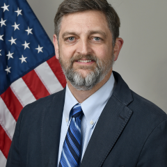 Man with a beard wearing suit and tie, smiling by an American flag.