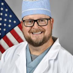 Smiling man in white lab coat, blue scrub top, and surgical cap with American flag behind him.