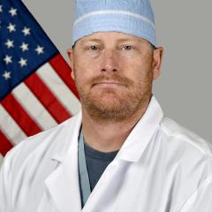 Male doctor in white lab coat and blue cap standing in front of the American flag.
