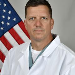 Man in white lab coat and green stethoscope stands before an American flag.