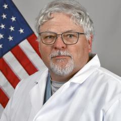 Man in white lab coat with grey hair and glasses standing before American flag.