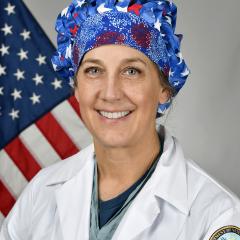 Female doctor in white lab coat and patriotic scrub cap standing in front of an American flag.
