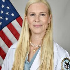 Blonde woman in white coat with US flag behind her.