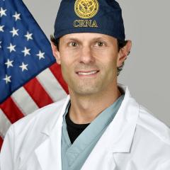 Man in white lab coat and blue cap standing in front of an American flag.