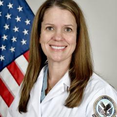 A smiling woman in a white coat stands in front of an American flag.