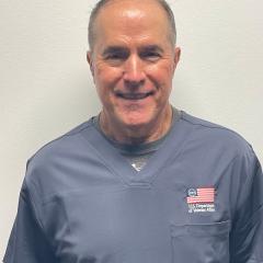 Smiling man in navy scrubs with US flag and text on chest pocket.