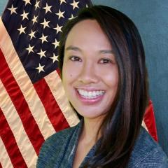 A smiling woman with long black hair stands in front of an American flag.