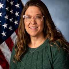 A smiling woman with long hair in a green blouse stands in front of an American flag.
