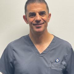 Middle-aged man in gray scrubs smiling against a plain white wall.