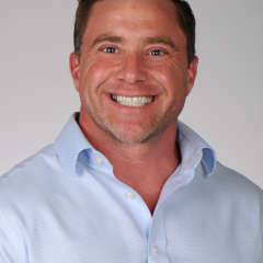 Smiling man in blue shirt with rolled up sleeves against plain white background.