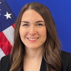 Portrait of woman with long brown hair smiling in front of American flag.