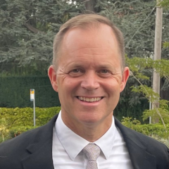 Man in suit and tie smiling in front of green bushes.
