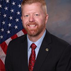 Man in suit and red tie standing before American flag.