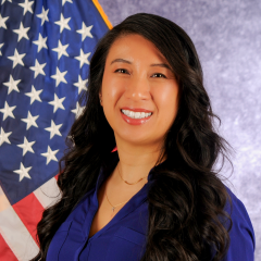 Woman with long dark hair wearing blue shirt smiling in front of American flag.