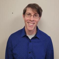 Smiling man in blue shirt and glasses stands against plain background.