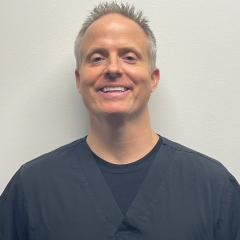 A smiling man in black scrubs stands against a white wall.