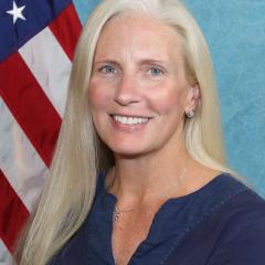 Blonde woman with blue shirt smiling in front of an American flag.