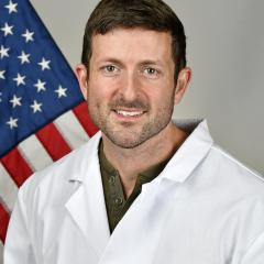 A man in a white lab coat smiles in front of an American flag.