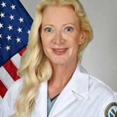 Portrait of a blonde woman in a white lab coat with an American flag in the background.