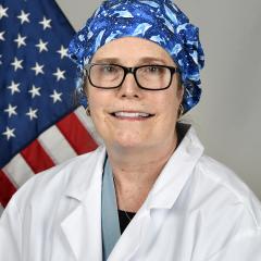 Woman in white lab coat and blue scrub cap smiling in front of American flag.