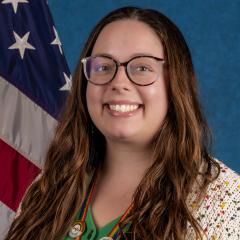 Woman with long hair, glasses, and medal smiling by flag.