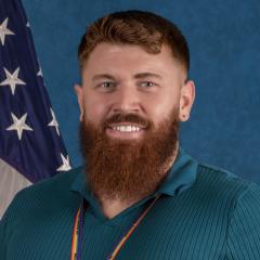 A smiling man with a red beard stands in front of an American flag.