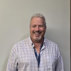 Man with gray hair and beard, wearing blue and white shirt, smiling at camera.