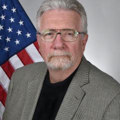 Man with white hair and beard wearing glasses and gray jacket, standing by American flag.
