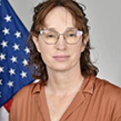 Woman in brown shirt with glasses stands before US flag.