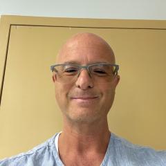 Portrait of bald man with glasses smiling in front of a yellow door.