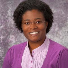 Woman with short curly hair wearing glasses, a purple cardigan, and pink shirt smiling.