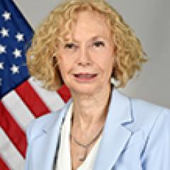 Blonde woman in blue blazer and white blouse with American flag in background.