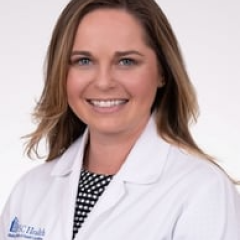 Female doctor with brown hair, smiling, wearing white lab coat with blue logo.