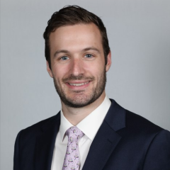 Man in suit and purple tie smiling against a gray background.