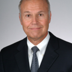 Headshot of a smiling man in a suit and tie against a gray background.