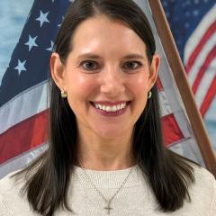 Woman with long black hair and light sweater, smiling in front of American flag.