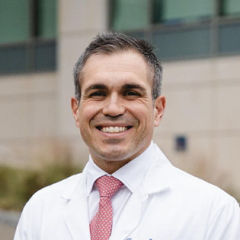 Smiling man in white lab coat and red tie outside building.