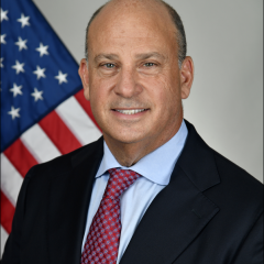 Smiling man in business attire with American flag in the background.
