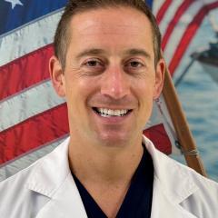 Smiling man in white lab coat with American flag in background.