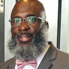 Bald man with gray beard wearing glasses, gray suit, and pink bow tie.