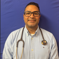 Smiling man in blue shirt with stethoscope around neck against blue backdrop.