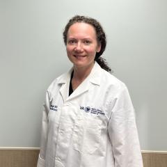 A smiling woman in a white lab coat stands against a beige wall.